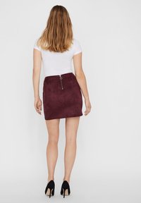 Saias mini em suede bordô com uma textura suave, apresentando um fecho de zip centralizado e um design de cintura alta. Sapatos de salto alto pretos completam o visual.