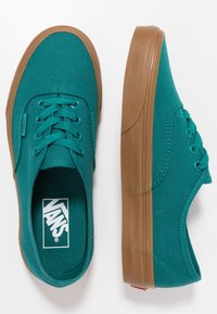 Baskets en toile bleu sarcelle avec semelles en caoutchouc marron, cinq œillets pour les lacets, et une étiquette logo sur le côté. Design plat avec texture lisse.