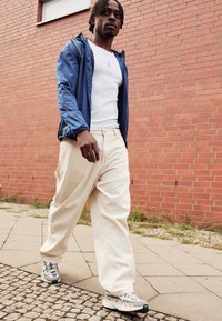 Blå vindjacka, vit linne topp, krämfärgade vida byxor och grå sneakers. Texturerad tyg och kontrasterande färgdetaljer. Tegelvägg i bakgrunden.