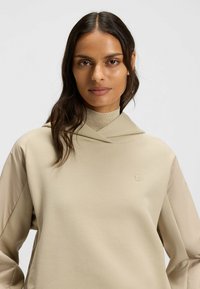 Felpa beige con colletto alto, orlo irregolare e maniche lunghe. Presenta un tessuto morbido con un accento di logo discreto.