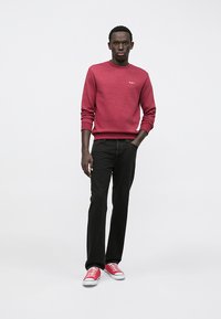 Maglione girocollo in pile rosso, jeans neri e sneakers in tela rossa. Il maglione presenta un piccolo logo sul petto. Design minimale.
