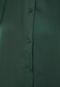 Ciemnozielona bluzka o gładkiej, błyszczącej teksturze z sześcioma dopasowanymi guzikami z przodu, prezentująca klasyczny fason na guziki.