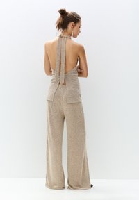 Vue arrière d'une combinaison sans manches en tissu beige scintillant, avec des jambes larges et un dos nu agrémenté de détails à nouer au cou et à la taille.