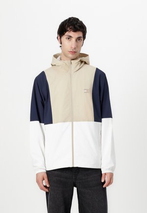 Color-blocked jacket featuring beige, navy, and white segments, lightweight material, front zipper, and a hood with elastic cuffs.