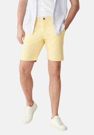 Homme portant un short jaune clair, des baskets blanches, un T-shirt blanc et une chemise à rayures bleu clair partiellement déboutonnée, les mains dans les poches.