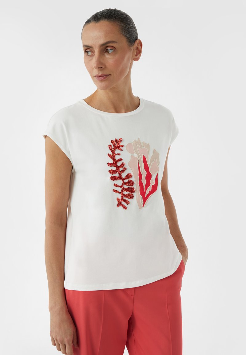 Witte top met korte mouwen, versierd met rode en beige bloemapplicaties. Gladde textuur met een ronde halslijn, gecombineerd met koraalkleurige broeken.