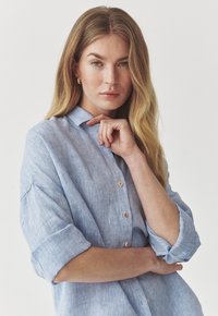 Camisa de lino azul claro con cuello, botones en el frente y mangas remangadas. Presenta una tela texturizada y un ajuste relajado. Sin estampados ni adornos.