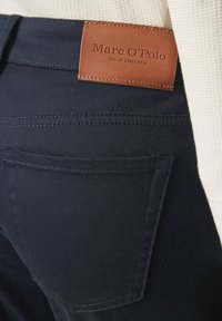 Pantaloni blu navy con una texture liscia e un'etichetta in pelle marrone con la scritta "Marc O'Polo" in vita, dettagli di cucitura visibili.