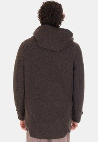 Persona con capelli ricci che indossa un cappotto con cappuccio a spina di pesce marrone scuro, in piedi di spalle contro uno sfondo semplice.