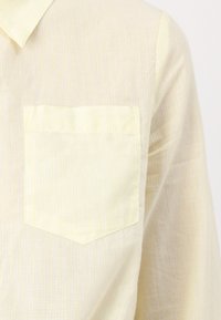 Chemise rayée jaune clair avec une poche. Tissu doux, col boutonné et surface subtilement texturée avec de fines lignes verticales.