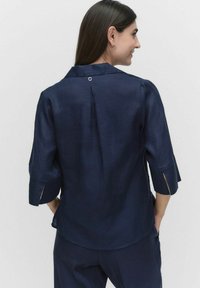 Mörkblå kortärmad blus med krage, med en veck bak och utskärningar på ärmarna. Slät textur, lättviktig tyg.