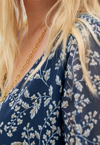 Námořnická květinová blůza se světle květinovými vzory, výstřihem ve tvaru V a poloprůhlednou tkaninou. Obsahuje zlatý řetízkový náhrdelník.