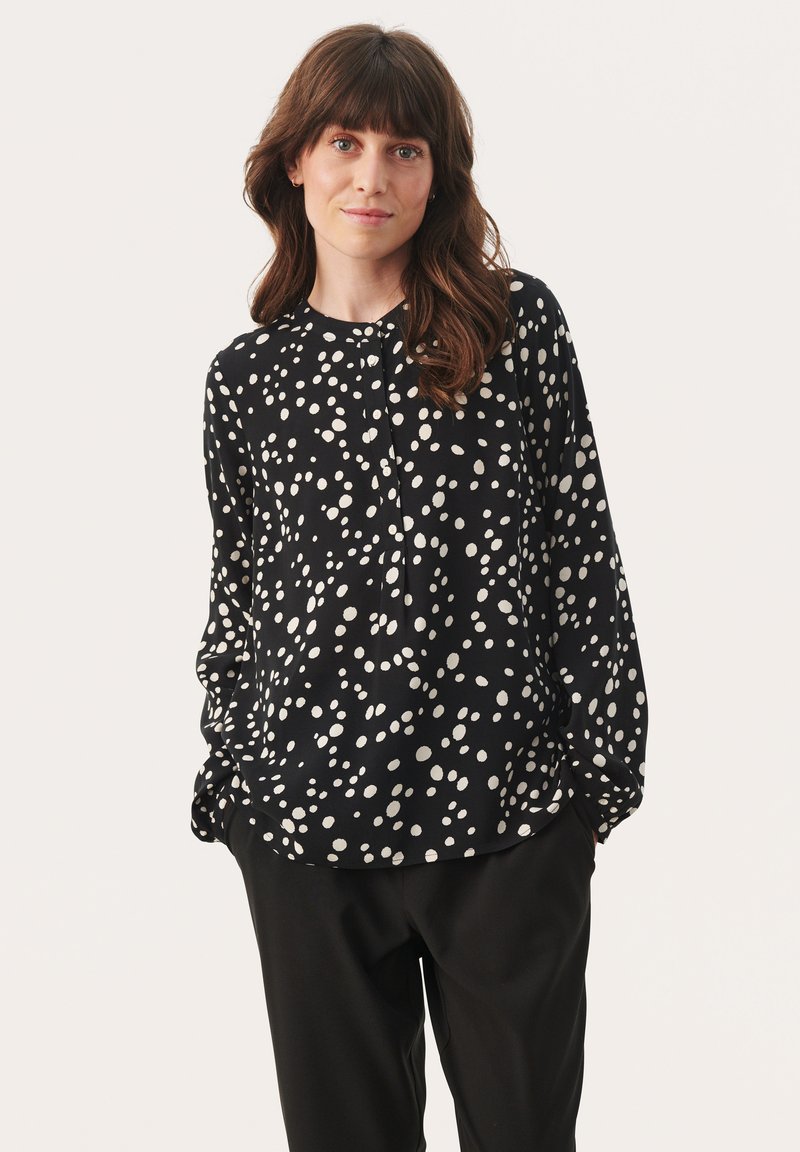 Zwarte blouse met lange mouwen en witte stippenpatroon, met een ronde halslijn en knopenrij, gecombineerd met zwarte broek.