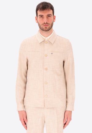 Uomo che indossa una giacca di velluto beige con chiusura a bottoni e pantaloni abbinati, in piedi davanti a uno sfondo chiaro e uniforme.