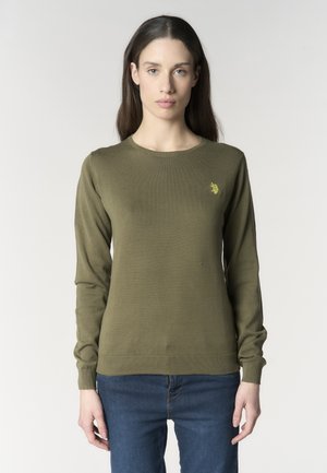 Giovane donna con capelli castani lunghi indossa un maglione verde oliva a maniche lunghe con un piccolo logo giallo e jeans blu, in posizione neutra.