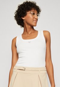 Hvid ribbet tanktop med en rund udskæring og lille broderet logo. Kombineret med en højtaljet beige nederdel med plisséer.