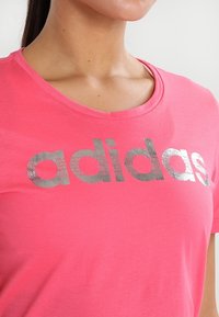 Žena v jasně růžovém tričku s lesklým stříbrným logem "adidas" vytištěným na hrudi.
