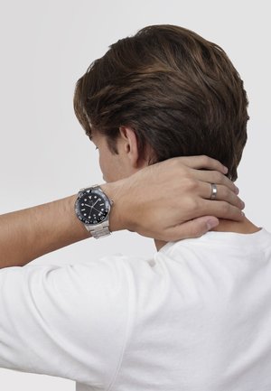 Junger Mann in weißem Hemd, der sich den Nacken berührt, trägt eine silberne Armbanduhr und einen silbernen Ring an seiner linken Hand.