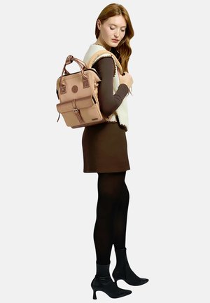 Frau trägt ein braunes Kleid, weiße Weste, schwarze Strumpfhosen und Stiefel und hat einen beigen Rucksack mit Fronttasche und zwei Tragegriffen über der Schulter.