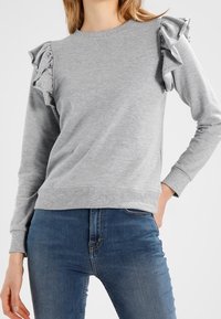 Vaaleanharmaa collegepaita, jossa on röyhelöyksityiskohtia hartiasaumoissa, pitkät hihat, resorit hihansuissa ja pyöreä pääntie. Päällä siniset farkut.