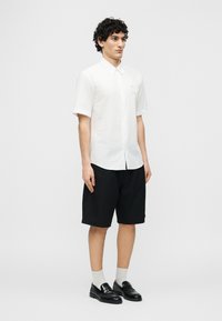 Νεαρός άνδρας με σγουρά μαλλιά, που φοράει λευκό πουκάμισο με κοντά μανίκια και κουμπιά, μαύρα σορτς μέχρι τα γόνατα, λευκές κάλτσες και μαύρα loafers, όρθιος.