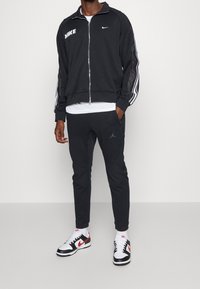 Chaqueta negra Nike con cremallera y logo blanco, mangas a rayas; pantalones jogger negros a juego; zapatos en blanco y negro con acentos rojos y calcetines blancos.