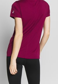 Camiseta deportiva color marrón con mangas cortas, textura suave, escote de cuello redondo, detalles en las costuras laterales y logo en la manga. Combinada con pantalones negros.