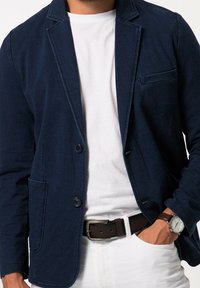 Marineblauwe blazer van zacht materiaal, met twee voorzakken, een enkele knoopsluiting, revers en contrasteert met een wit shirt en riem.