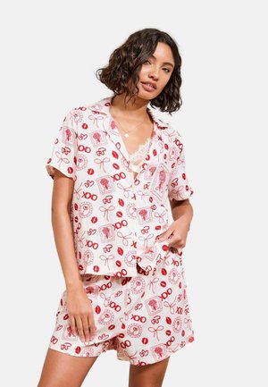 Vrouw draagt witte korte mouw pyjama set met rode kus-, strik- en "XOXO"-patronen, met daaronder een spaghettibandjes top met kanten randje zichtbaar.