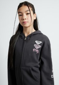 Zwarte zip-hoodie met een voorzak, met kleurrijke hartontwerpen en "Roxy"-tekst. Zacht stof, casual fit en lange mouwen.