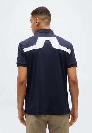 Moški, ki nosi mornarsko modro polo majico z belo kotno črto na zgornjem delu hrbta, oblečen v bež hlače, obrnjeno stran od kamere.