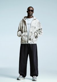 Man in zwarte zonnebril, witte hoodie met grafisch ontwerp, zwarte wijde broek en grijze sneakers staat met zijn handen in zijn zakken tegen een lichte achtergrond.