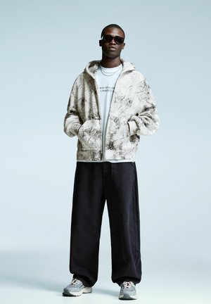 Man wearing black sunglasses, white graphic hoodie, black wide pants, and gray sneakers standing against a light background with hands in pockets.