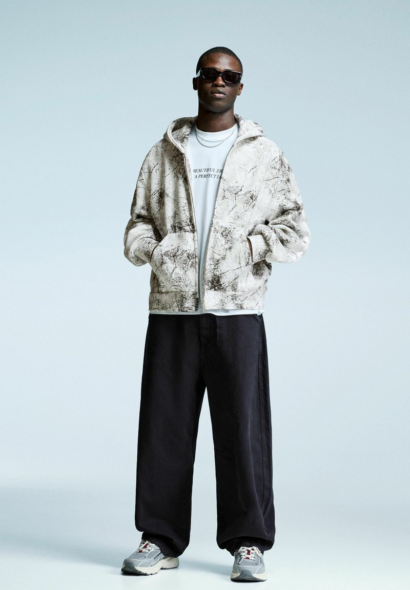 Man in zwarte zonnebril, witte hoodie met grafisch ontwerp, zwarte wijde broek en grijze sneakers staat met zijn handen in zijn zakken tegen een lichte achtergrond.
