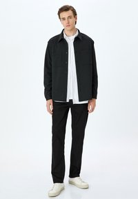 Camicia nera a bottoni con due tasche sul petto, indossata sopra una camicia bianca, abbinata a pantaloni neri e sneakers bianche, su uno sfondo semplice.