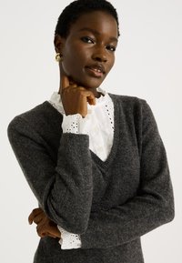 Suéter de lana gris con escote en V, superpuesto sobre una camisa blanca con cuello y borde de encaje, que presenta un acabado texturizado y mangas largas.