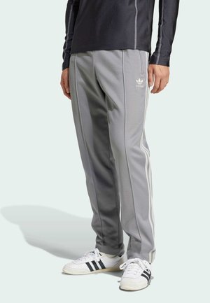 Mężczyzna w szarych spodniach dresowych Adidas z białymi paskami i białymi butami Adidas z czarnymi paskami, stojący na jasnym tle.