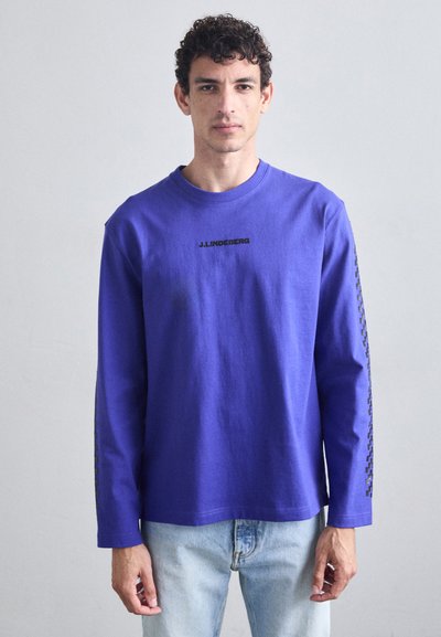 T-shirt violet à manches longues en coton avec un col rond et un logo noir sur la poitrine ; présente un motif à carreaux sur les manches.