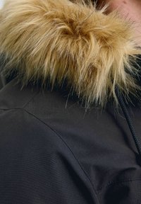 Sort jakke med en lækker, gyldent-brown plys faux fur-hette. Har et struktureret stof og synlige syninger langs skuldersømmen.