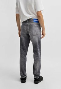 Grå denimjeans med lys vask og lige ben, der har et blåt HUGO-label i taljen og på baglommerne, båret med sorte sko.