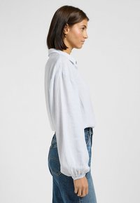 Camicia button-up a strisce bianco e azzurro, con colletto, vestibilità ampia e maniche lunghe a sbuffo, abbinata a jeans in denim blu a vita alta.