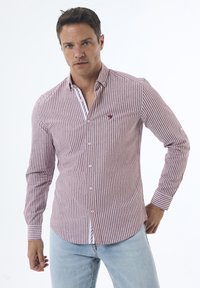 Camisa de botones de manga larga a rayas rojas y blancas con cuello, que presenta un pequeño logo en el pecho y puños abotonados. Llevada con jeans de color azul claro.