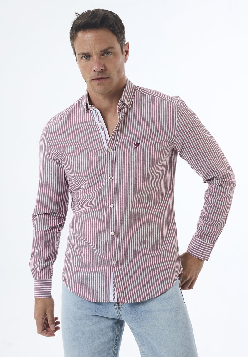 Camisa de botones de manga larga a rayas rojas y blancas con cuello, que presenta un pequeño logo en el pecho y puños abotonados. Llevada con jeans de color azul claro.