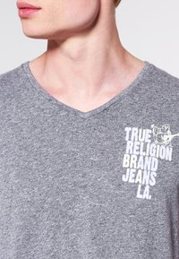 Graues V-Ausschnitt-T-Shirt aus weichem Stoff, mit einem weißen Logotext auf der linken Brustseite. Lässiges Design mit strukturiertem Finish.