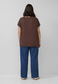 Persoon met schouderlang donker haar draagt een losse bruine top, een blauwe wijde jeans en witte sneakers, staand met het gezicht naar een effen grijze muur.