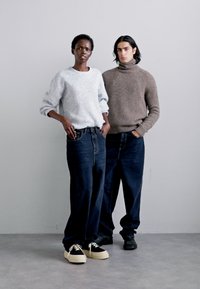Two models stand together. The first wears a gray sweater and dark, loose-fitting jeans; the second wears a brown turtleneck and similar jeans.