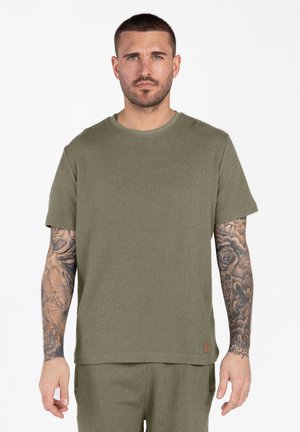 Mann mit kurzen Haaren und Tätowierungen auf beiden Armen, trägt ein olivgrünes, strukturiertes kurzärmeliges Hemd und passende Shorts, steht.