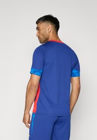 Maglietta sportiva da uomo di colore blu con accenti rossi, maniche blu texture e design aderente. Il materiale sembra liscio con modelli a contrasto.