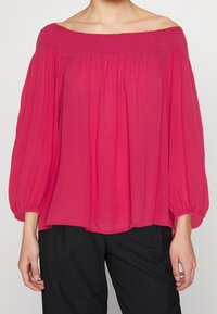 Blusa off-the-shoulder di un vivace rosa, con scollatura arricciata e maniche ampie e fluttuanti con polsini elastici. Tessuto leggero e fluido.