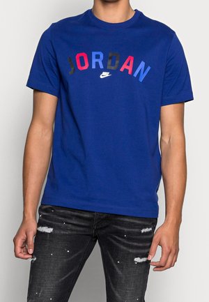 Niebieski bawełniany t-shirt z nadrukiem graficznym z napisem "JORDAN" w kolorach czerwonym, niebieskim i czarnym. Krótkie rękawy i okrągły dekolt. Połączony z czarnymi, postrzępionymi jeansami.
